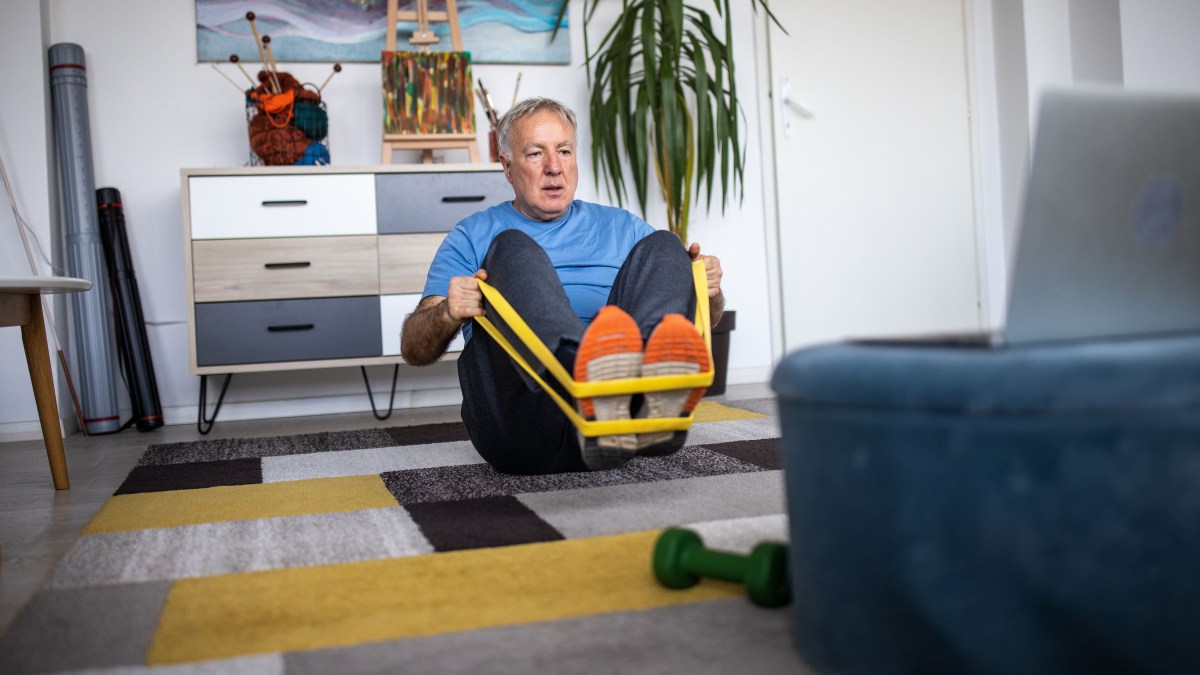 An older man exercises at home.  He was sitting on the carpet, facing the screen of a laptop that was open.  He uses a band of resistance, wrapped around his feet, and pulls towards it.  There is a small green pumpkin near him.