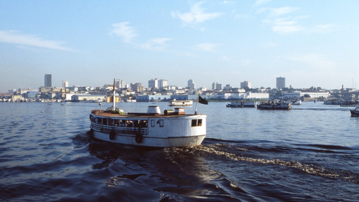 In this picture you see a white ferry full of tourists traveling on the Amazon River.  In the background you can see the horizon of the city of Manaus, Brazil.